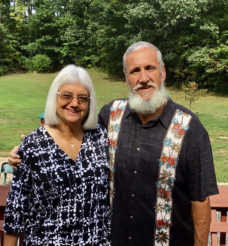 Deb and her husband, Rolland, smile with a backdrop of green grass and pine trees.