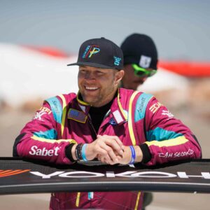 Don Wickstrum, the Fastest Pastor and cancer survivor, smiles and wears racing uniform while leaning on the rear fin of a race car. 